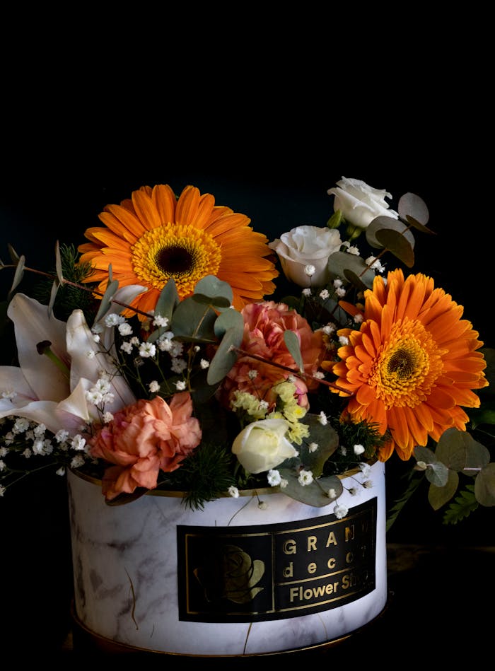 A beautiful floral arrangement with orange gerberas, roses, and eucalyptus in a marble vase against a dark background.